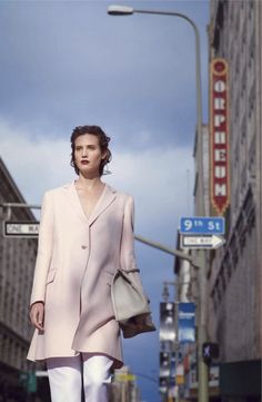 a woman is walking down the street in a pink coat and white pants with heels