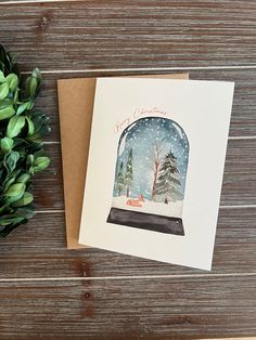 a snow globe christmas card on top of a wooden table next to a potted plant