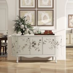 a white cabinet with flowers on it in front of three framed pictures and a table