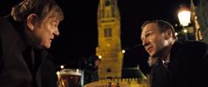 two men sitting at a table in front of a tall building with a clock tower