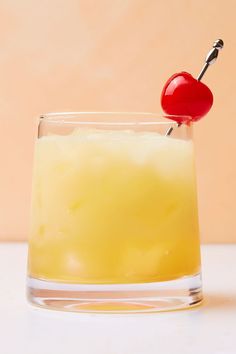 a glass filled with yellow liquid and a cherry on the top, sitting on a table