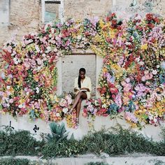 a woman sitting in front of a wall covered with flowers and greenery on the outside