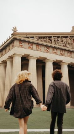 two people holding hands in front of a building with columns and statues on the side