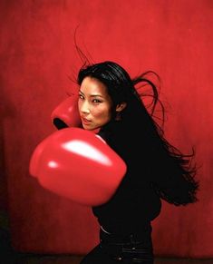 a woman with long black hair and red boxing gloves is posing for a photo in front of a red wall