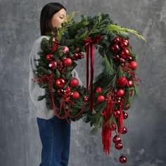 a woman holding a christmas wreath with red balls and greenery on the front, while standing against a gray background