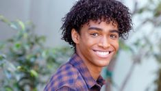 a young man with curly hair smiles at the camera while standing in front of a bush