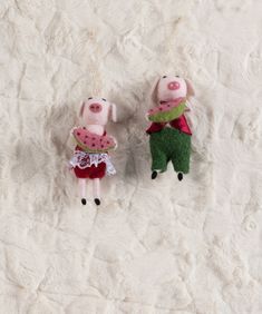 two stuffed pigs hanging from strings on a white furnishing surface with one pig wearing a watermelon outfit and the other in a green dress