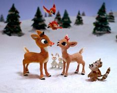 there are many toy deers in the snow with birds flying above them and trees behind them