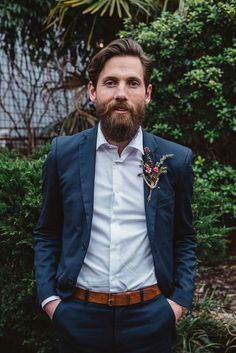 a man with a beard wearing a suit and flower boutonniere standing in front of bushes