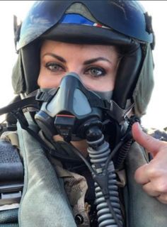 a woman wearing a gas mask gives the thumbs up sign with her right hand as she sits in an aircraft cockpit