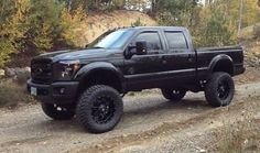 a black truck is parked on the side of a dirt road with trees in the background