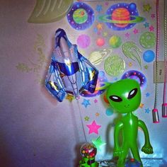 a green alien toy sitting on top of a table next to a wall covered in stars and planets