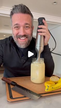 a man holding a blender in his right hand and smiling at the camera with bananas around him