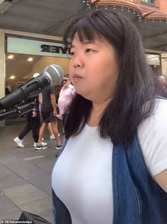 a woman standing next to a microphone in front of a building with people walking by