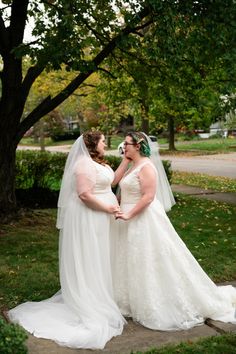 two women in wedding gowns standing next to each other on the sidewalk near trees