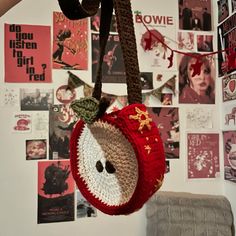 a crocheted apple hanging from a wall with posters on the wall behind it