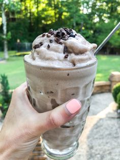 a person holding up a drink with ice cream and chocolate chips on it in front of a park