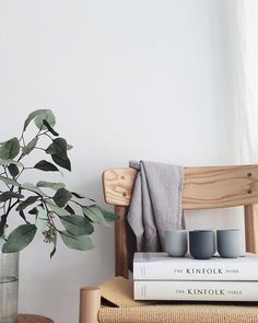 a wooden chair sitting next to a vase filled with green leaves and two cups on top of each other