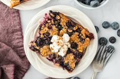 two plates with blueberry crumble cake on them next to silverware and berries