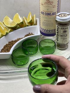 a person holding a glass with green liquid in front of some lemons and spices