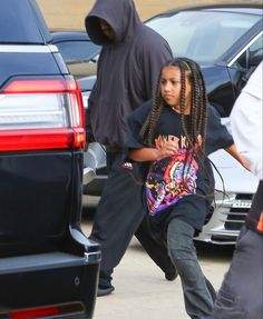 a person in a black hoodie is walking by some cars and another person with dreadlocks