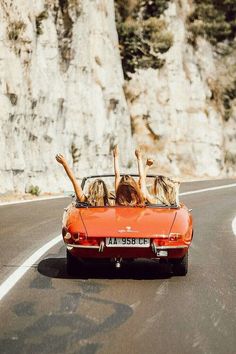 two people in an orange convertible driving down the road with their arms up and legs out