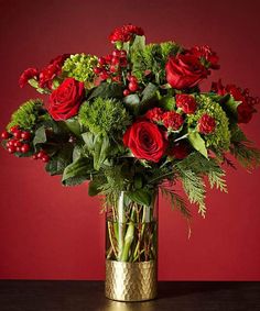 a vase filled with red roses and greenery on top of a wooden table next to a red wall