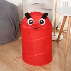 a red plastic trash can with eyes on it's face sitting on the floor next to a bed