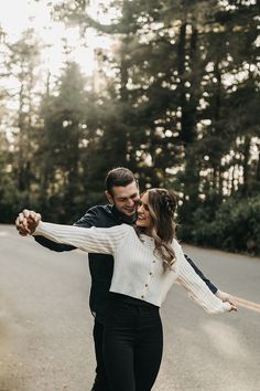 a man and woman dancing on the road in front of some trees with their arms around each other