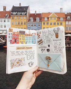 someone is holding up an open book in front of some buildings and boats on the water