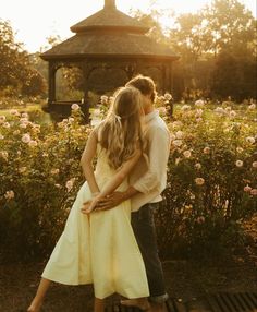 a man and woman are hugging in front of some bushes with flowers behind them, while the sun is setting