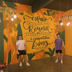 two men standing in front of a colorful wall with words on it and lights hanging from the ceiling