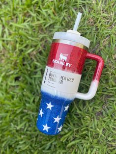 a red, white and blue coffee cup sitting on top of green grass with the word stanley printed on it