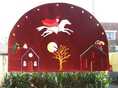 a large red sign with horses and houses painted on it's sides in front of some bushes