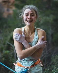 a woman is smiling and holding her arms around her chest while standing in the woods