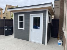 a small gray shed with a white door and window on the side of it, next to a black trash can