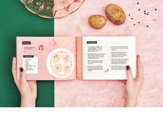 two hands holding an open book on top of a pink and green tablecloth with food