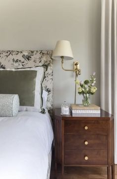 a bed with white sheets and pillows next to a lamp on a night stand in front of a window