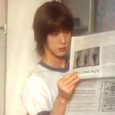 a young man reading a newspaper while standing in front of a wall with his hair pulled back