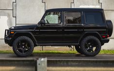 a black mercedes benz g - class is parked in front of a concrete wall and grass