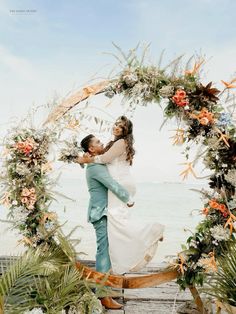 a bride and groom standing in front of an archway with flowers on the beach at their wedding