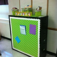 a green and white bulletin board with notes on it, next to a black filing cabinet