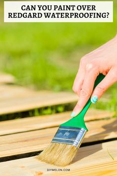 a person holding a paint brush on top of a wooden deck with the words can you paint over redcard waterproofing?