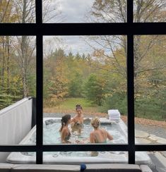 three people sitting in a hot tub with trees outside the window and one person standing by it