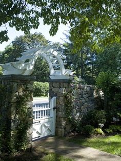 a white gate surrounded by stone walls and greenery on the side of a road