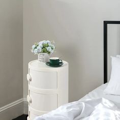 a white nightstand with flowers and a cup on it next to a bed in a room