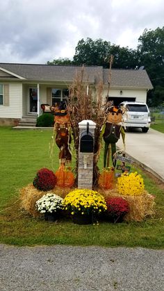 a mailbox surrounded by flowers and hay in front of a house with a car parked on the driveway