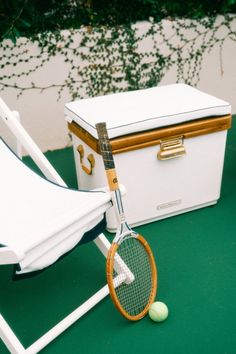 Vintage-style wooden tennis racket leaning on a white deck chair next to a luxury outdoor cooler, showcasing timeless recreational charm.