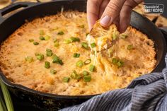 a person dipping cheese into a skillet filled with green onions and cheesy bread