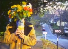 a man holding a vase with flowers on his head while standing next to a street